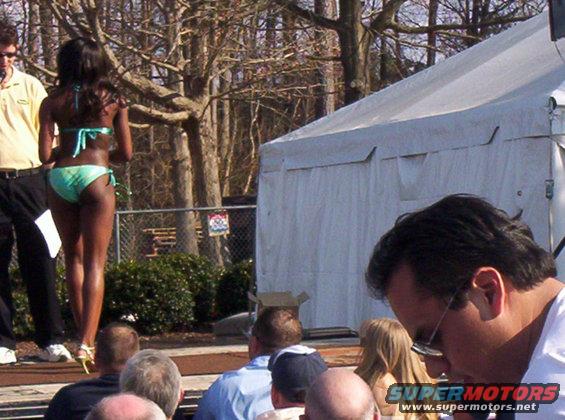 Woman in a teal bikini on a stage with trees and a tent in the background