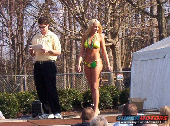 Woman in green and yellow bikini on stage with a man holding papers, outdoor event