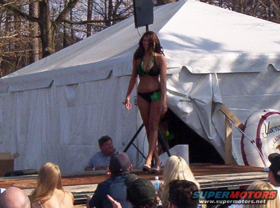 Woman in bikini on outdoor stage with tent behind her and audience in foreground