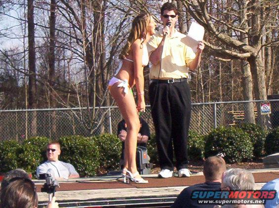 Man with microphone talks to woman in bikini on stage, audience watches