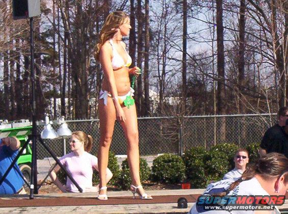 Woman in white and yellow bikini on a stage with trees and a fence in the background