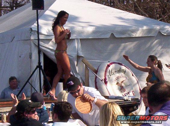 People gathered around a table at an outdoor event, with a woman in a bikini on a stage behind them