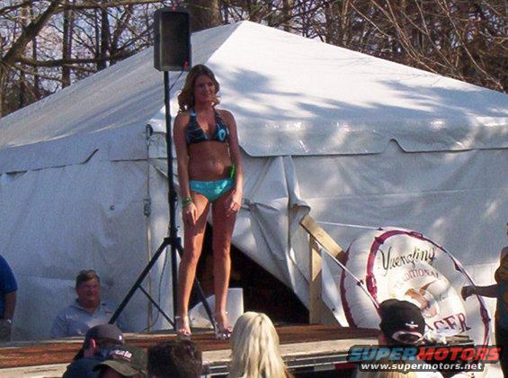 Woman in bikini on stage in front of a tent, with a large circular sign behind her
