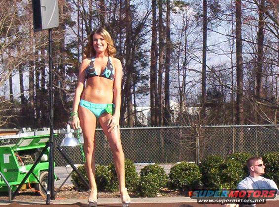 Woman in a bikini stands on a stage with trees and a fence in the background