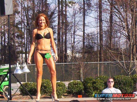 Woman in black bikini with shamrock decoration on stage, trees and fence in background