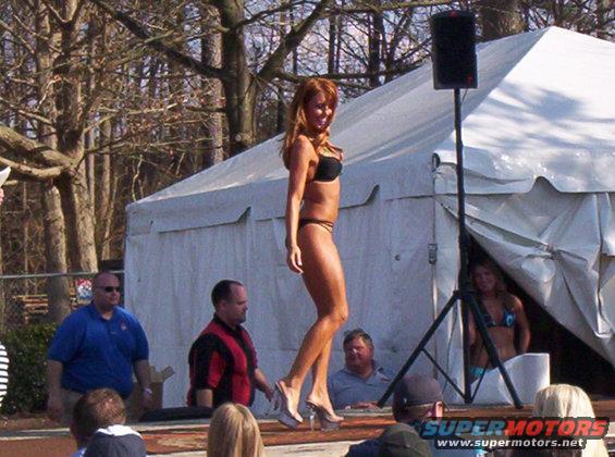 Woman in bikini walking on stage, tent and trees in background