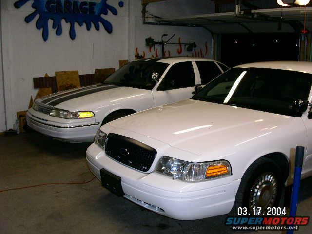 Two white Ford Crown Victoria police cars parked in a garage. One has black racing stripes on the hood