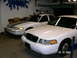 2001 Ford Crown Victoria - Two white Ford Crown Victoria police cars are parked side-by-side in a garage, with one racing stripes on its hood.