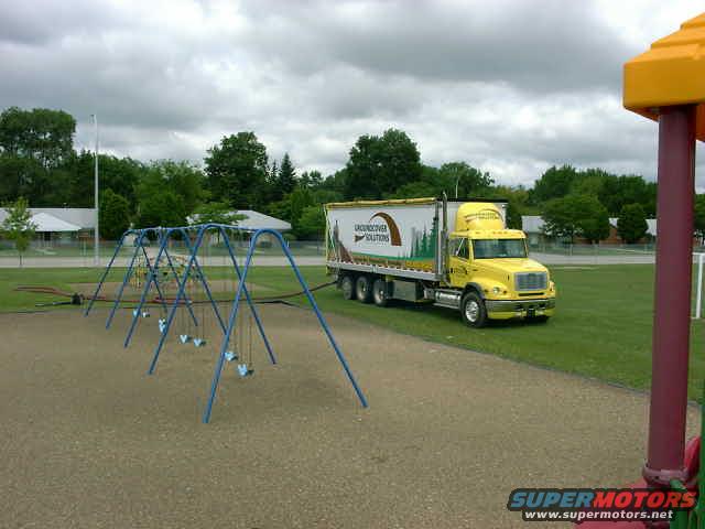 Yellow semi-truck with a white trailer parked on grass near a playground