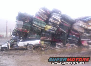 Pile of crushed cars, one white pickup truck in foreground, muddy ground, overcast sky