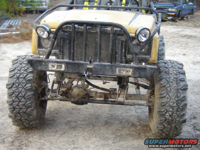 Front view of a yellow off-road buggy with large tires and a roll cage. A blue car is visible in the background