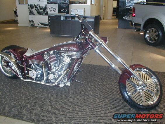 Maroon custom motorcycle with chrome accents and a large front wheel, parked indoors on a patterned carpet