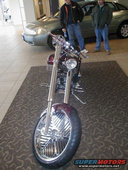 Custom chopper motorcycle with chrome accents parked on a rug, two men and a car in the background