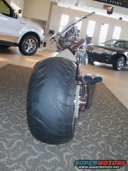 Custom motorcycle with a wide rear tire and chrome accents, parked indoors next to a white truck and a black sedan
