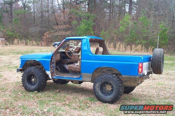 1987 Ford Bronco with doors removed, lifted suspension, and large off-road tires. Spare tire mounted on rear. Interior visible