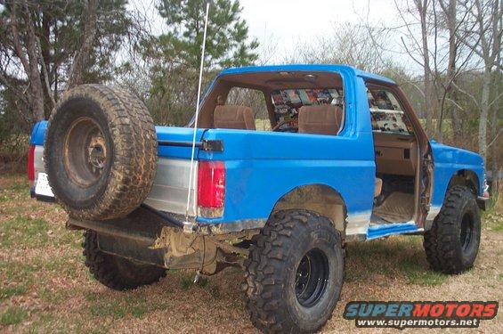 Blue Ford Bronco with a spare tire on the back, lifted with large tires, and the driver's door removed