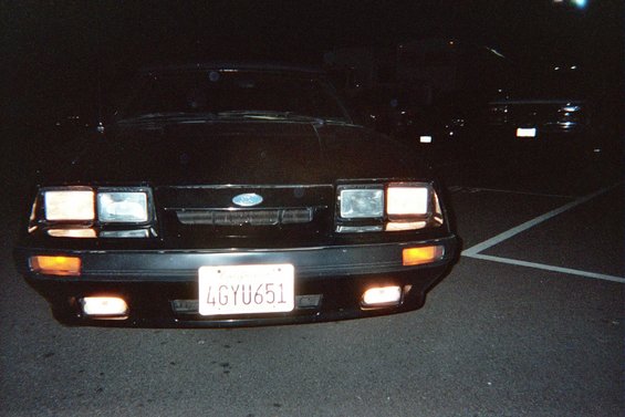 Front view of a black 1985 Ford Mustang with its headlights on. Another vehicle is visible behind it