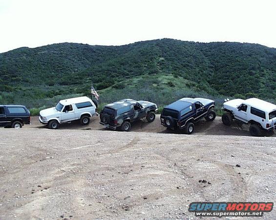 Five Ford Broncos parked on a dirt road with a hilly, green background. An American flag is visible