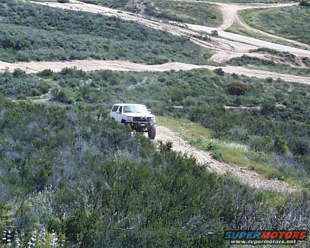 1993 Ford Bronco driving on a dirt track through a brushy, hilly area