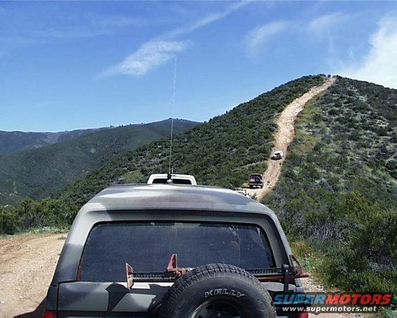 Camouflage SUV rear view with spare tire and jack, driving on a dirt road up a hill with other vehicles