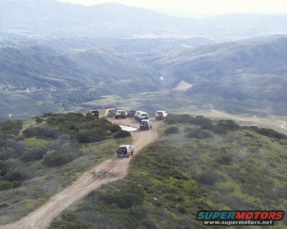 Several SUVs drive on a dusty mountain road, with a town visible in the distant valley