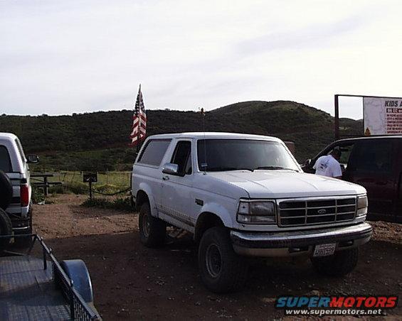 White 1992 Ford Bronco parked on dirt lot with American flag and hills in background. Another vehicle is partially visible on the right