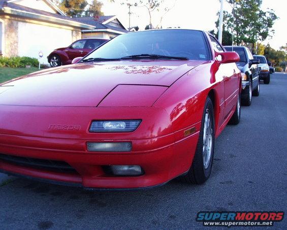 Red Mazda RX-7 parked on street, front quarter view. Other cars are behind it