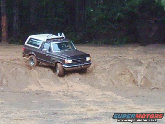 Dark gray Ford Bronco with white top driving on a dirt hill, trees in background