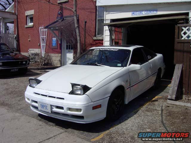 White coupe with pop-up headlights parked in front of a garage. A dark car is parked to the left