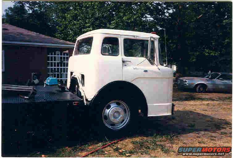 White cab-over truck on a flatbed trailer, front quarter view. Another car is visible in the background