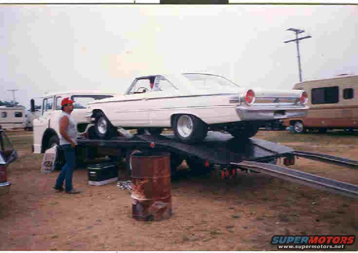 1957 Ford Fairlane on car hauler, man standing nearby, RV and truck in background