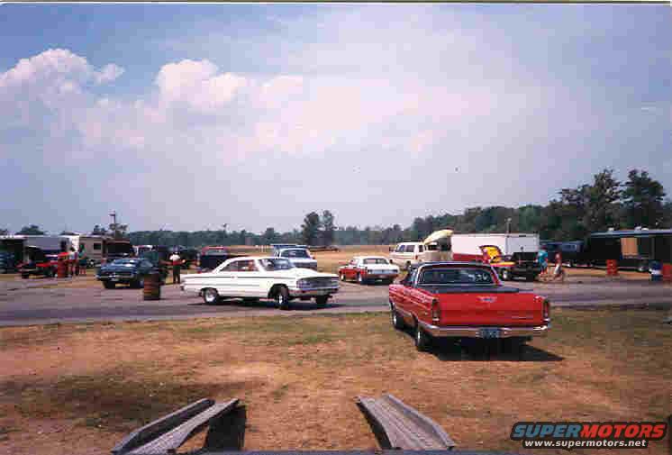 drag strip with a white sedan, red pickup, and other vehicles parked around