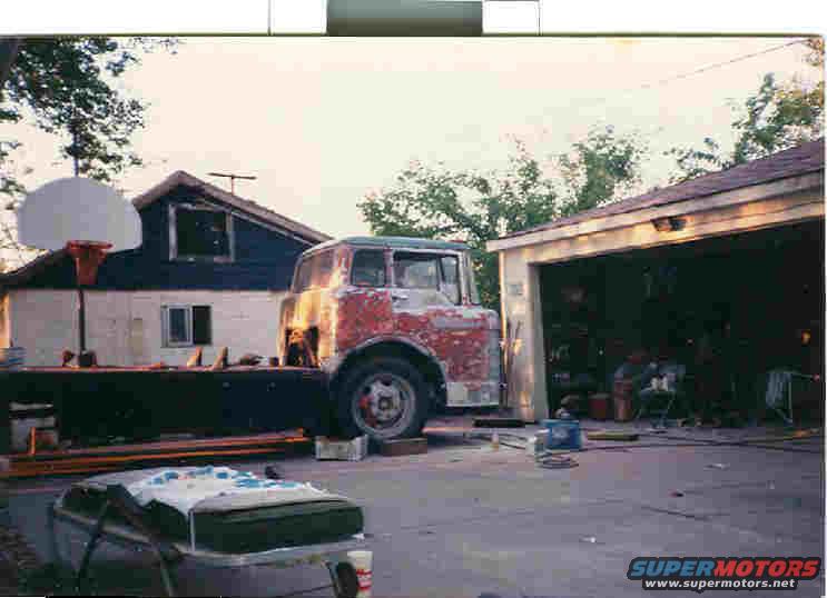 Truck cab with peeling red paint, sitting on blocks next to a garage. A basketball hoop is visible behind it