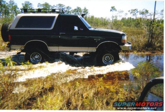 Black and tan Ford Bronco driving through shallow water, with a spare tire on the back and a roof rack