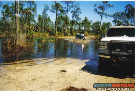 Two trucks driving through a shallow body of water with trees in the background