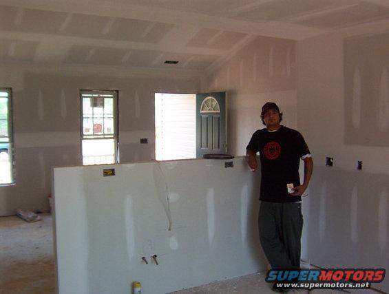 Man in black t-shirt and cap leans against drywall in a room under construction