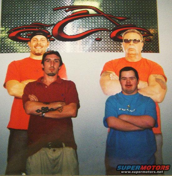 Four men stand with arms crossed in front of a metallic wall with a red motorcycle logo. The man on the far left wears an orange t-shirt and a cap. The man next to him wears a maroon polo shirt. The man on the far right wears an orange t-shirt and sung...