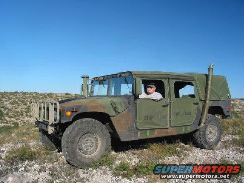 Humvee with a person driving, parked on a rocky, dry landscape under a clear blue sky