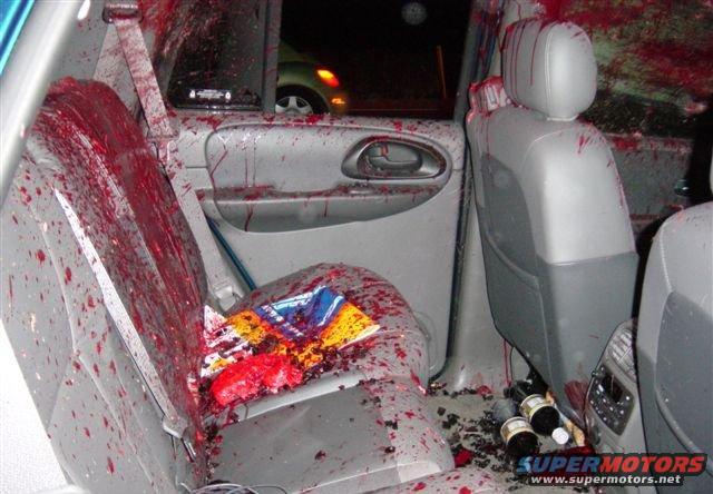 Interior of a vehicle with seats and doors covered in blood splatters. Bottles and debris are on the floor