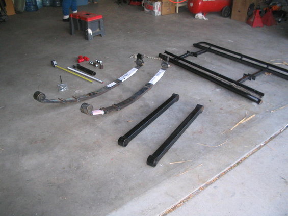 Leaf springs and black metal bars on concrete floor. Tape measure and red fittings nearby