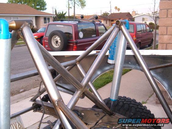 Custom roll cage and suspension parts on a vehicle. A red SUV with a spare tire on the back and a red van are visible in the background