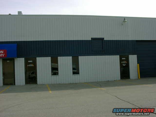 Building with blue and white siding, dark blue stripe, and multiple doors and windows. A yellow pole is visible near a large dark garage door