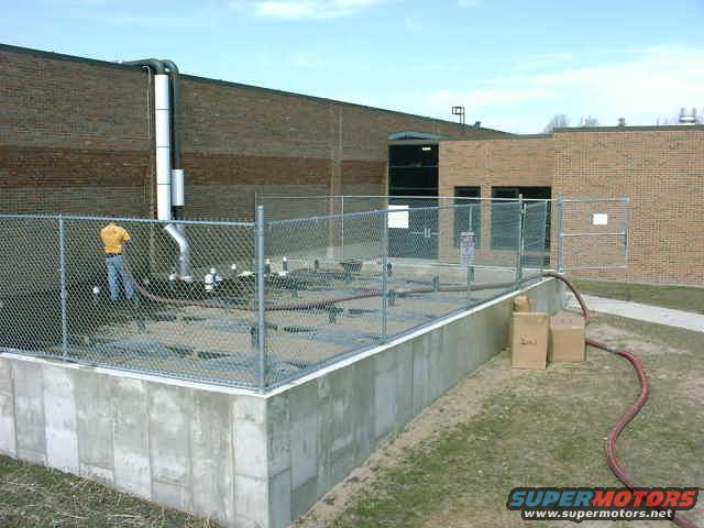 Industrial building with a chain-link fence enclosing pipes and a person. Two cardboard boxes are on the grass