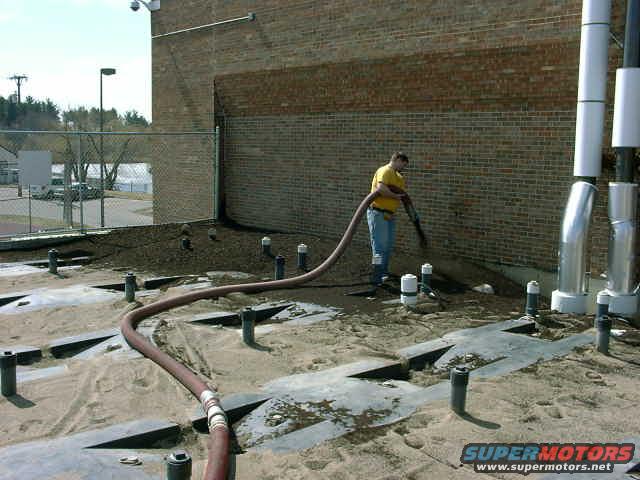Worker using a hose on a construction site with a brick building and pipes