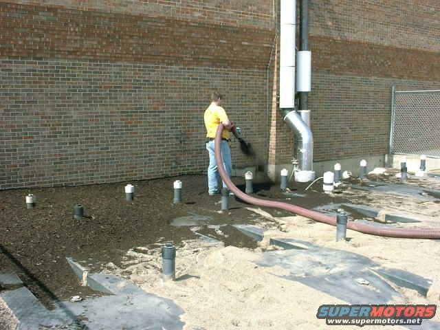 Worker in yellow shirt using a hose near a brick building with pipes and ground pipes