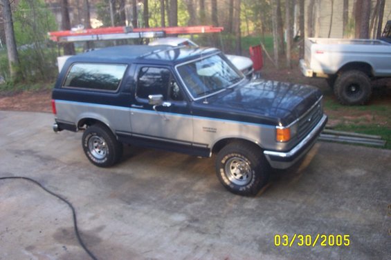 Two-tone blue and silver Ford Bronco with a soft top, parked on concrete. Another lifted truck is visible in the background