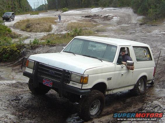 White 1989 Ford Bronco navigating muddy off-road terrain with another SUV in the background