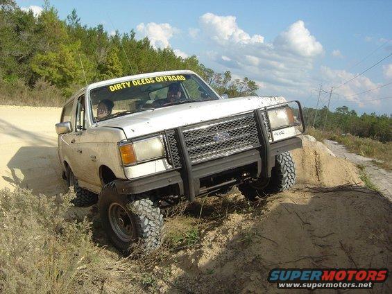 White SUV with 'DIRTY DEEDS OFFROAD' banner on windshield, driving on a dirt hill. It has a brush guard and off-road tires