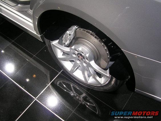 Close-up of a silver car's front wheel, showing the multi-spoke rim, brake caliper with 'V' logo, and perforated rotor. The car is on a black tiled floor