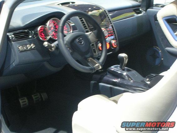 Car interior with dark blue dashboard, red-lit gauges, and white seats. A "SUPERMOTORS" logo is visible
